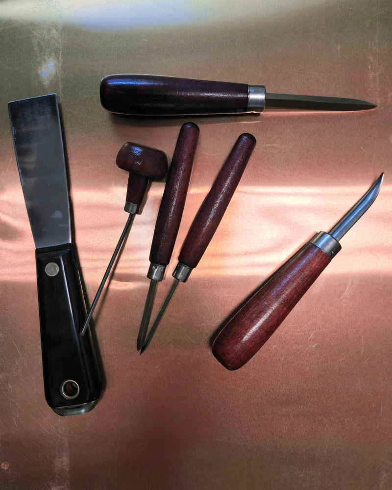 Dark wooden-handled printmaking tools arranged on a copper surface