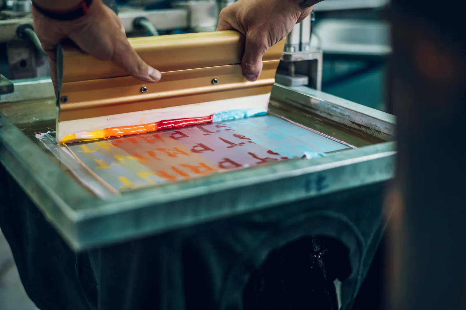 Hands pulling a squeegee loaded with colorful ink across a silk screen